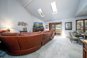 Ein Wohnzimmer mit braunem Ledersofa, Fernseher und Kamin. Es gibt Dachfenster, einen Essbereich und einen Blick ins Freie.