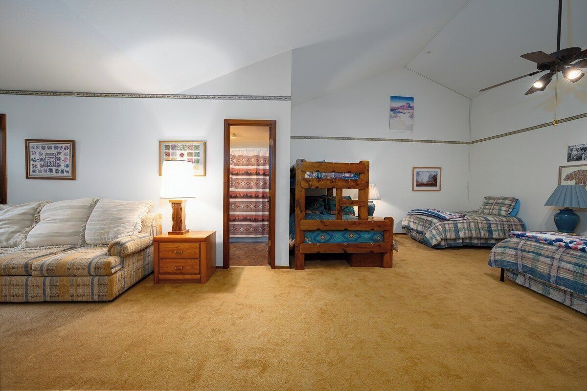 A bedroom with a sofa, two beds, a wooden chair, a nightstand, a lamp, and a ceiling fan.