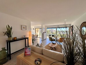 A modern living room with a sofa, coffee table, and potted plants. There is a dining area with chairs and a table, and a balcony with a view. - Marina view apartment, terrace & pool (Mandelieu-la-Napoule)