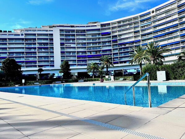 A modern hotel with a large swimming pool, multiple balconies, and palm trees. - Marina view apartment, terrace & pool (Mandelieu-la-Napoule)