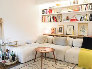 Un salon comprenant un canapé d’angle, une petite table ronde, une bibliothèque et un chariot à boissons avec des bouteilles.