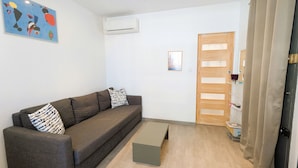Une pièce comprenant un canapé, une petite table, un appareil de climatisation, une porte en bois et une étagère avec divers objets.