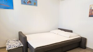 Une petite chambre avec un lit simple, un canapé-lit gris, une petite table de chevet avec un coussin à motifs et deux tableaux abstraits accrochés aux murs.