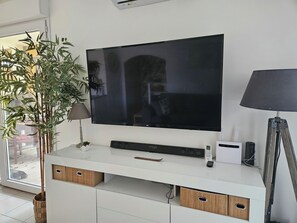 A modern living room with a large flat-screen TV mounted on a white console, a bamboo plant, a lamp, and a tripod on the right. - Apartment with terrace and garden in Audenge (Audenge)
