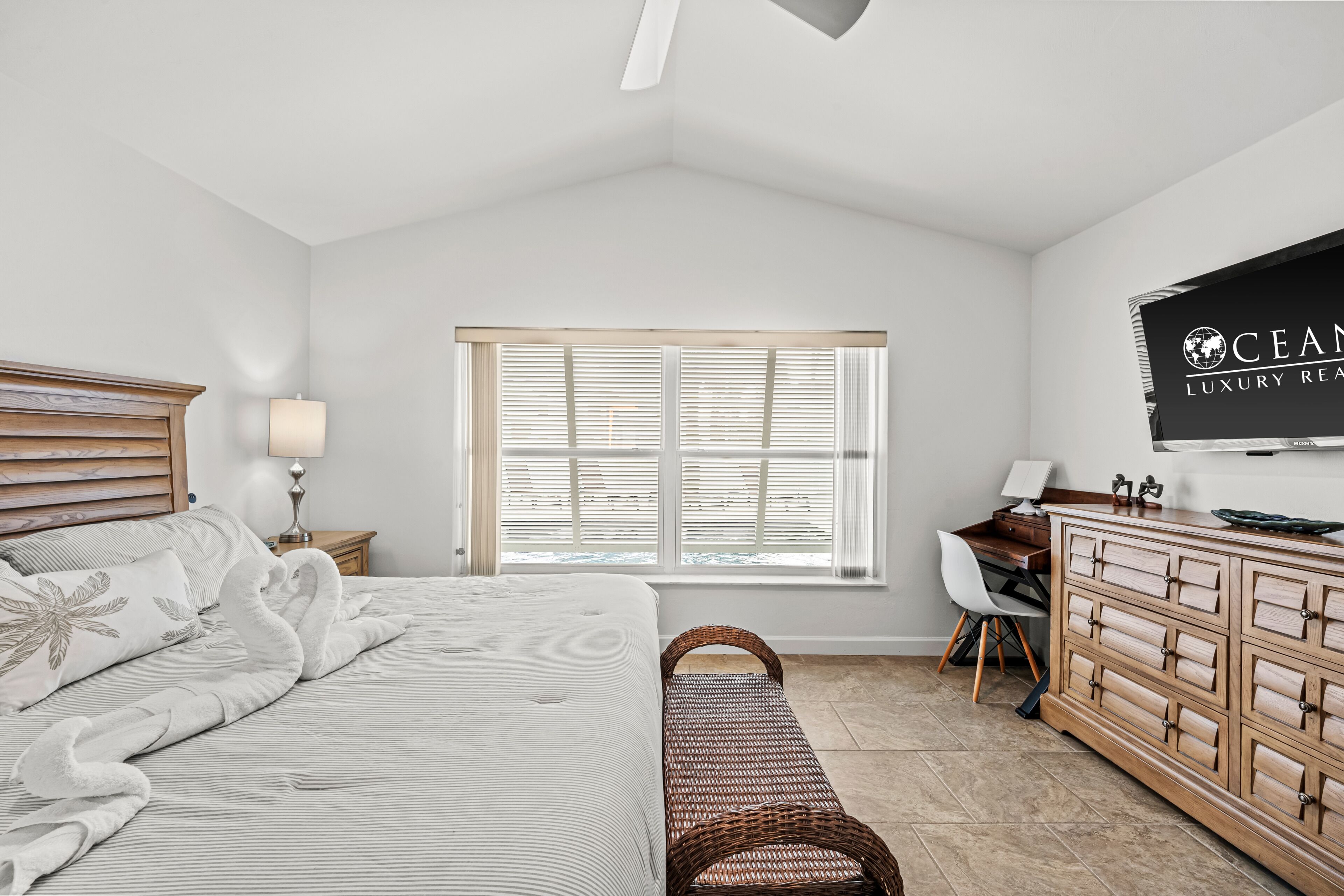 Une chambre à coucher comprenant un lit, un bureau, une commode, une fenêtre avec des stores et un téléviseur fixé au mur.