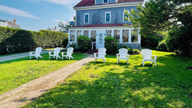 A large two-story house with a porch, surrounded by a well-maintained lawn and several white Adirondack chairs. - Historic 7-bedroom home in the heart of downtown in private setting,Pet friendly (New Shoreham)