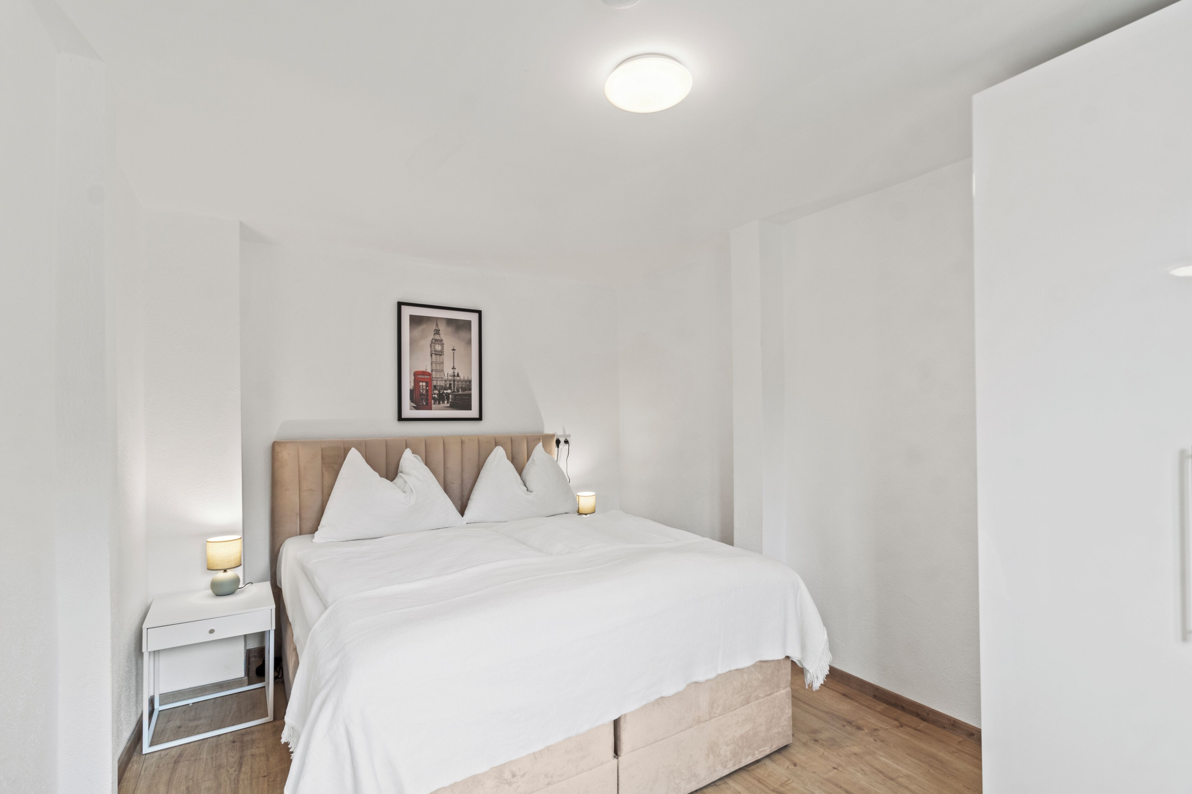 A bedroom with a bed, bedside tables, a nightstand, a wall-mounted picture, and a ceiling light.