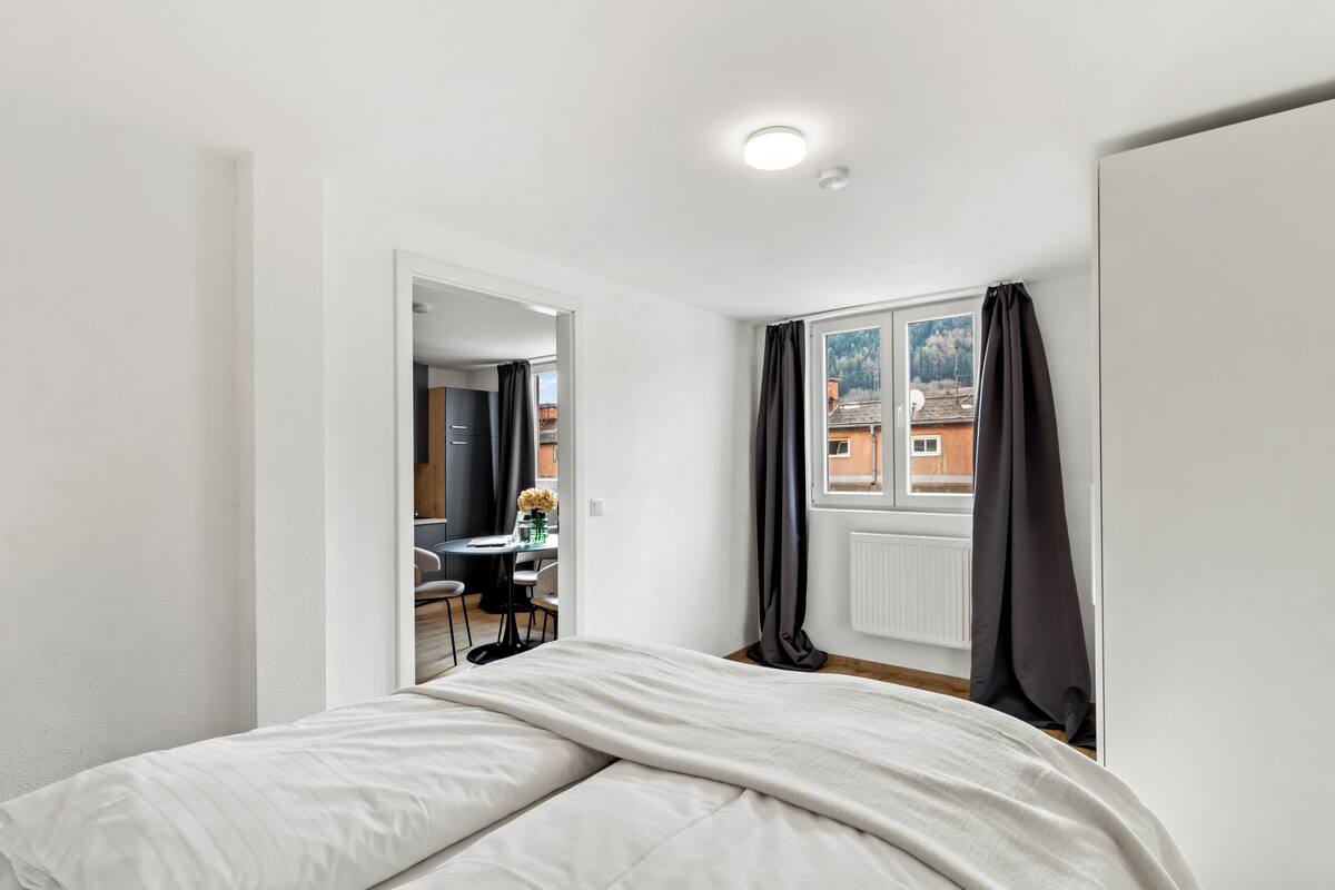 A bedroom with a bed, a window with curtains, a radiator, and a view of buildings outside.