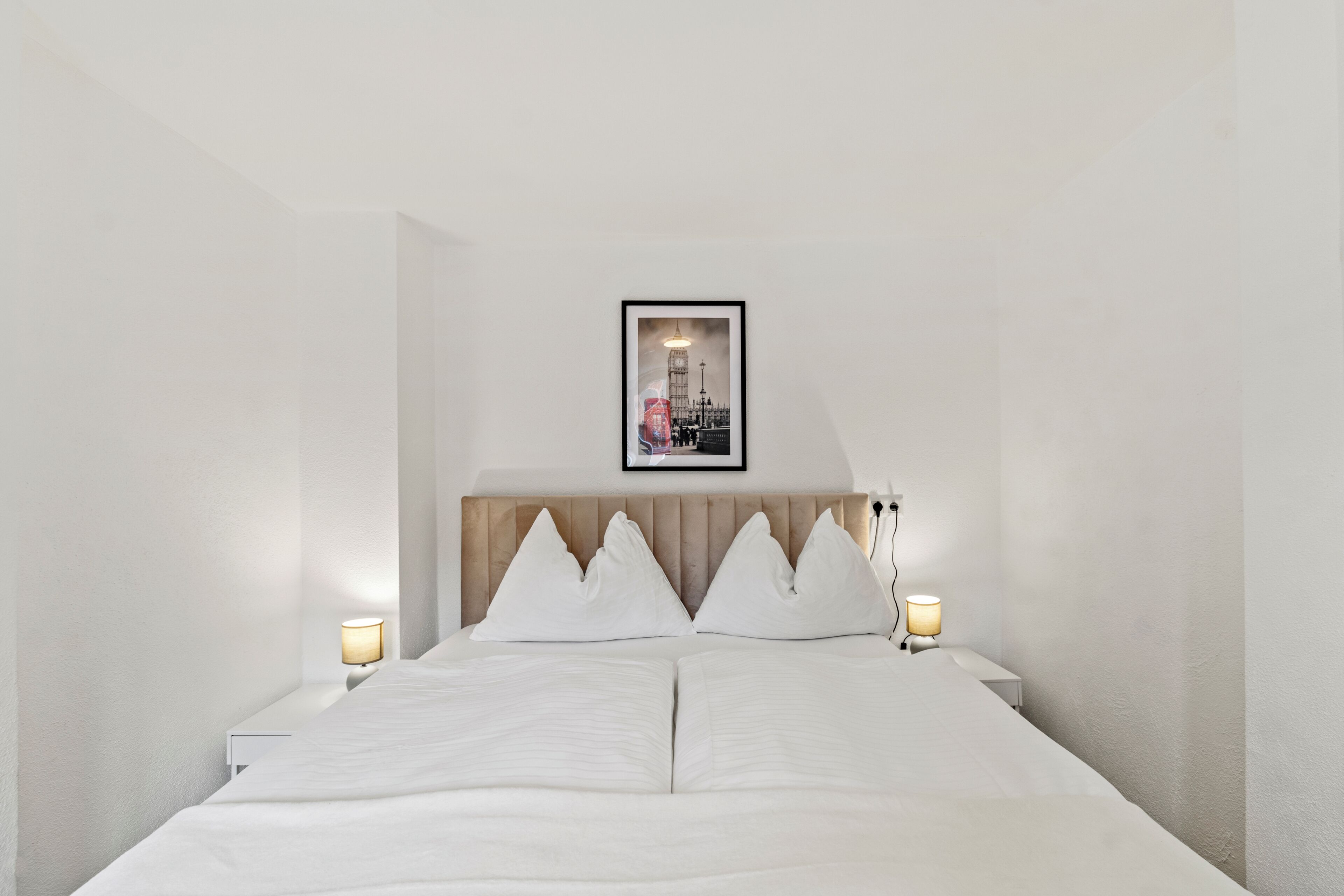 Een net opgemaakt bed met een hoofdbord, twee bedlampen en een ingelijst schilderij aan de muur.