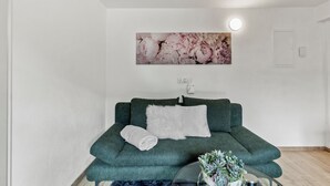 Ein Wohnzimmer mit einem grünen Sofa, weißen Kissen, einem aufgerollten weißen Handtuch, einem runden Tisch mit einer Pflanze und einem floralen Gemälde an der Wand.