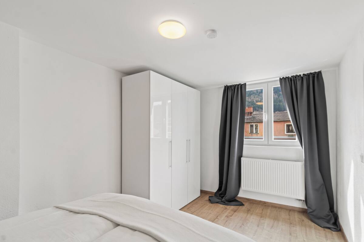 A bedroom with a bed, white wardrobe, window with curtains, and a view of buildings.