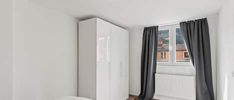 A bedroom with a bed, white wardrobe, window with curtains, and a view of buildings.