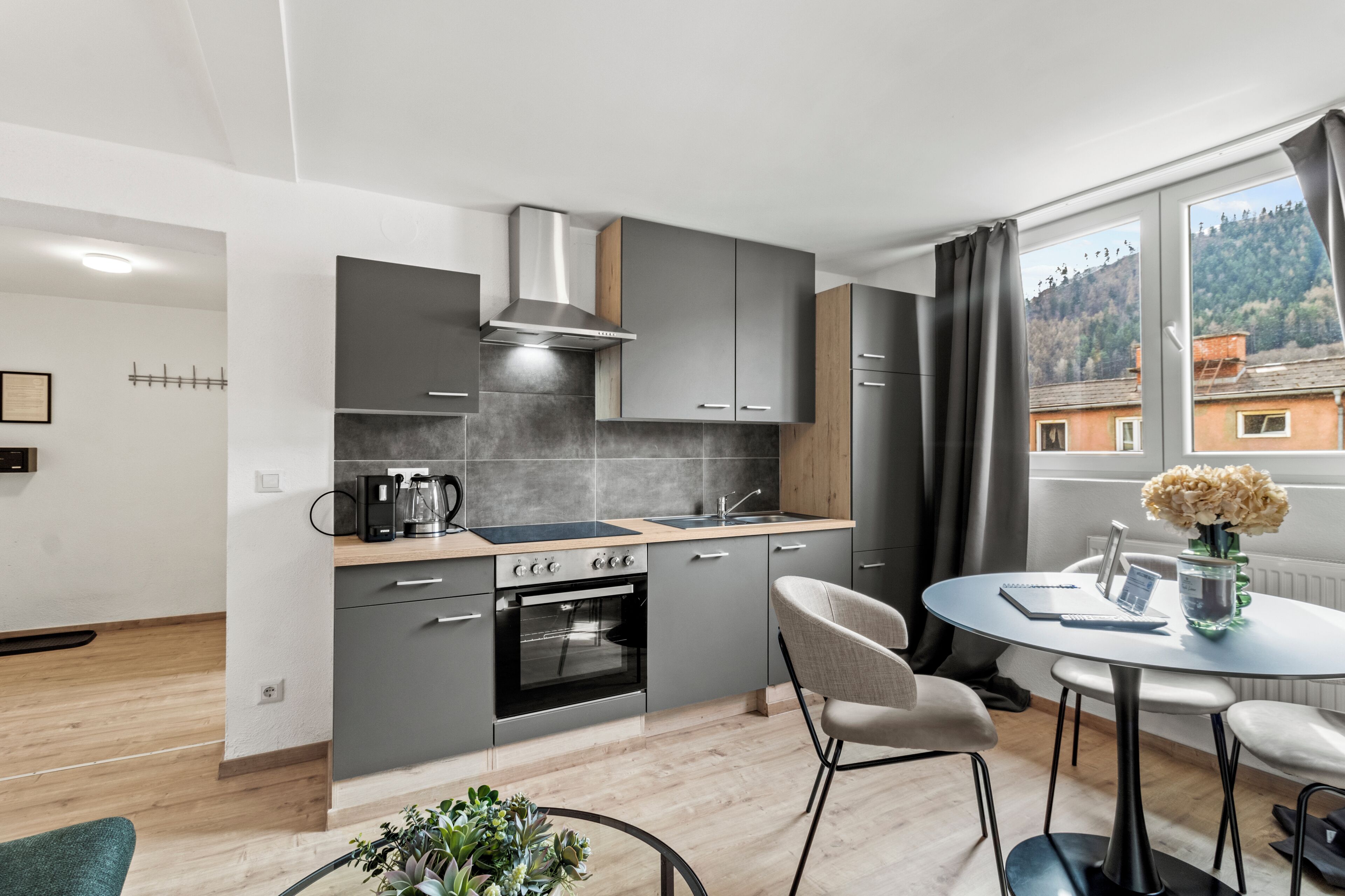 A modern kitchen with grey cabinets, a stove, and a sink. There is a dining area with a round table and chairs, and a window with a view of buildings.