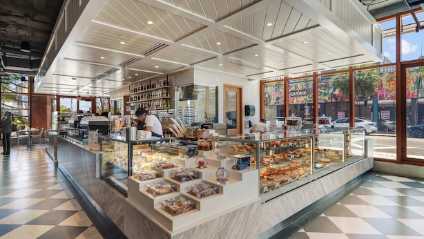 A bakery counter with various baked goods, a seating area with tables and chairs, and a view of the street outside. - 1BR + Balcony | Skyline Spa | Walk to Wynwood Walls (Miami)