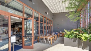 An outdoor cafe with tables and chairs, a glass door entrance, and a plant in the foreground. - 1BR + Balcony | Skyline Spa | Walk to Wynwood Walls (Miami)