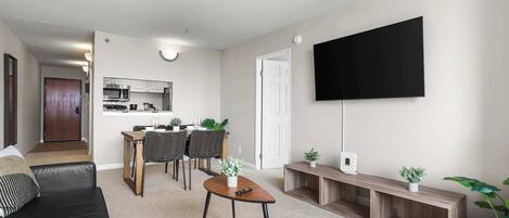 Een moderne woonkamer met een flatscreen-tv, een eethoek met een tafel en stoelen, en een keuken op de achtergrond.