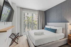 Ein Hotelzimmer mit Bett, Wandfernseher, Fenster mit Vorhängen und Balkon mit Pflanzen.