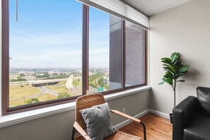 A room with a large window overlooking a highway and cityscape, a wicker chair with a cushion, and a potted plant. - Arlington Gem Stylish Apt with Pool and Gym (Arlington)