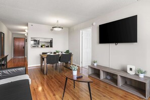 Ein modernes Wohnzimmer mit Flachbildfernseher, ein Essbereich mit Holztisch und Stühlen, eine Küche mit Mikrowelle und Schränken sowie ein Flur, der zu einer Tür führt.