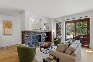 A modern living room with a stone fireplace, a sofa set, and a large window with a view of trees. - Tremblant l Paradise | Ski in & Ski out | Spa&Pool (Mont-Tremblant)