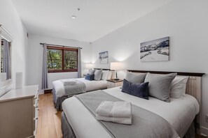 A hotel room with two beds, a window with curtains, a bedside table, and a wall-mounted lamp. - Tremblant l Paradise | Ski in & Ski out | Spa&Pool (Mont-Tremblant)