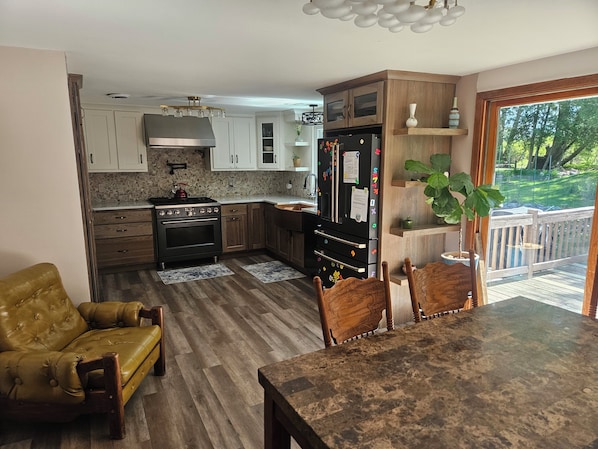 A kitchen with a stove, oven, refrigerator, and wooden cabinets. There is a dining table with chairs and a yellow armchair.