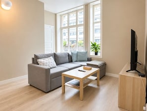 A modern living room with a grey sofa, a wooden coffee table, a TV on a stand, and a window with a view of buildings and greenery. - A40 - 2BR City Center - Cozy Apartment (Bergen)