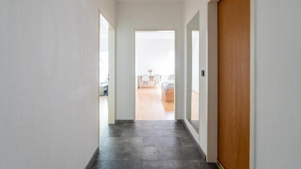 Un couloir avec une porte en bois, une vue sur une pièce où l’on aperçoit un lit et une table, et un sol carrelé.