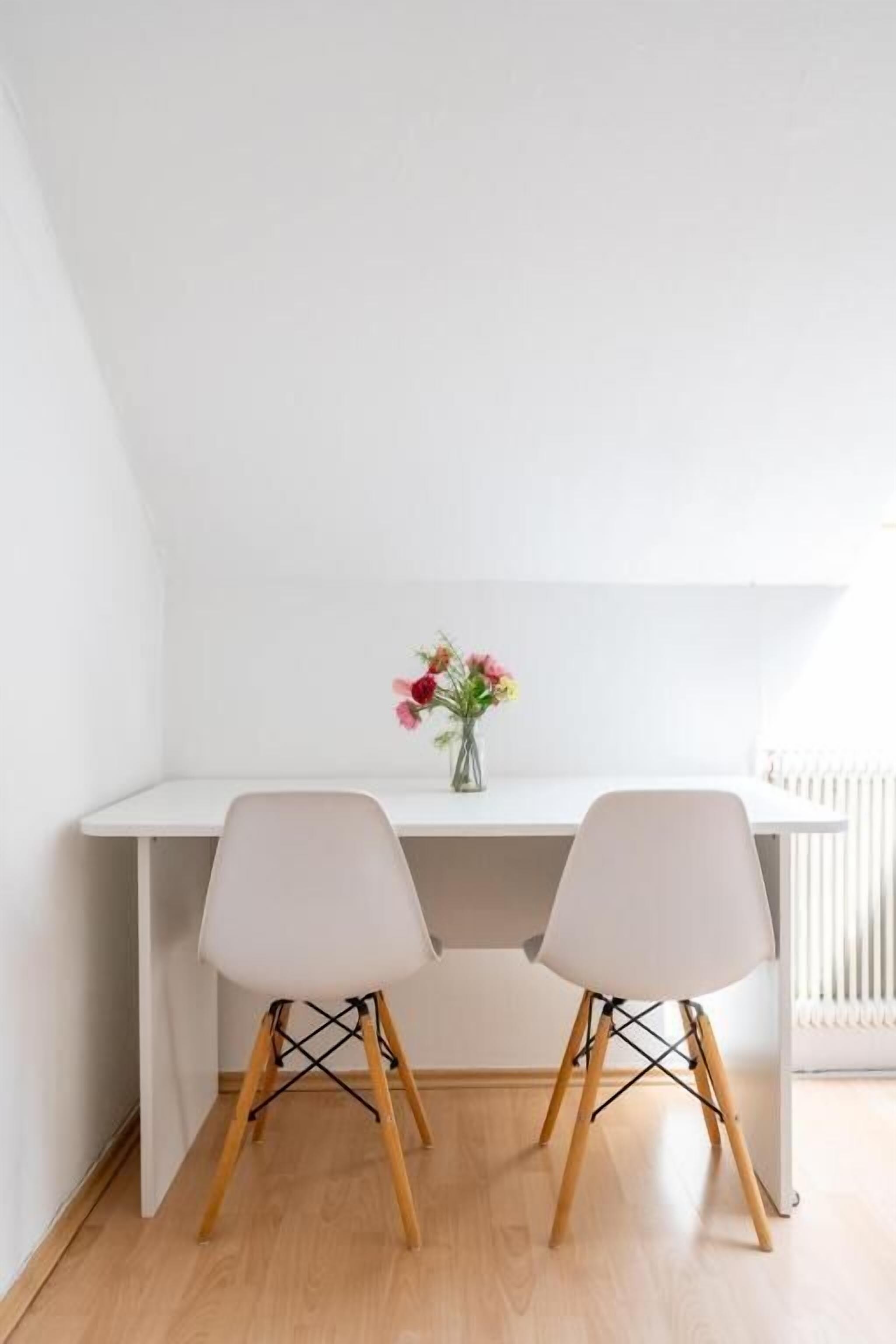 A white desk with two chairs, a vase with flowers, and a radiator in the background.