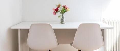 Un escritorio blanco con dos sillas, un florero con flores y un radiador al fondo.