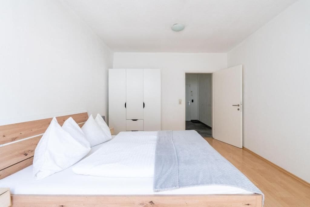 A minimalist bedroom with a wooden headboard, a bed with white bedding, and a grey throw.