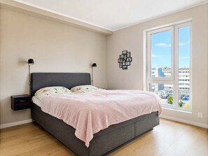 Un dormitorio con cama, mesitas de noche, ventana con vista a la ciudad y un cuadro colgado en la pared.
