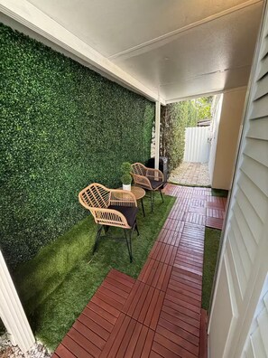 Een overdekt terras met kunstgras, houten terrasplanken en rieten meubilair.