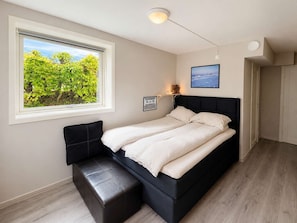 Un dormitorio con cama, un banco, una ventana con vista a follaje y una lámpara de pared, además de un cuadro enmarcado.