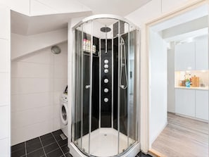 Un baño moderno con ducha acristalada, lavadora y una zona de cocina visible a través de una puerta abierta.
