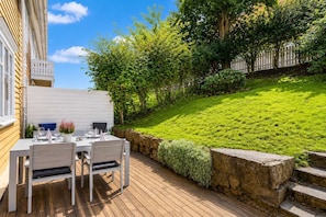 Une terrasse en bois avec une table dressée pour le repas, entourée d’un mur de pierre et d’une végétation luxuriante.