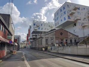 Une rue bordée d’immeubles, dont un bâtiment blanc doté de balcons et un bâtiment en briques avec une enseigne.