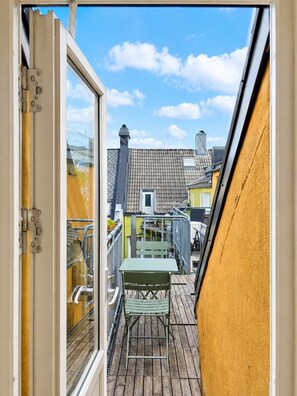 Un étroit balcon extérieur doté d’un plancher en bois, d’une rambarde en métal, ainsi que d’une table et d’une chaise vertes.