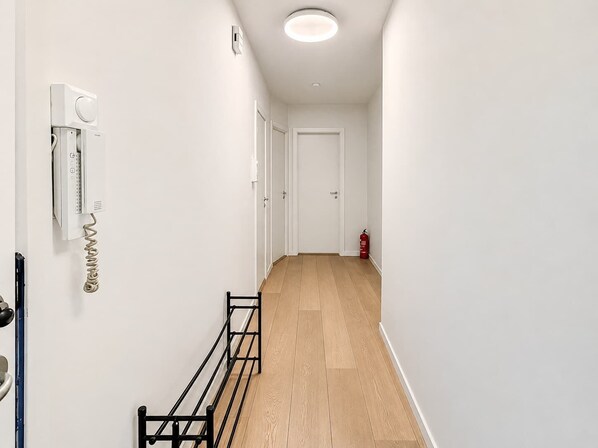 Un pasillo con pared blanca, un banco de metal negro, una puerta blanca al final y un extintor rojo a la derecha.