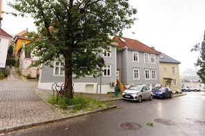 En boligvej med huse, et træ, parkerede biler og en brostensbelagt sti.