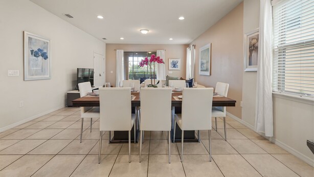 A dining area with a table set for four, chairs, a TV, and artwork on the walls. - EV_1800 - 6BR / 5BA Luxury Villa w/ Private Pool & Spa, Game Room, Resort Access (Kissimee)