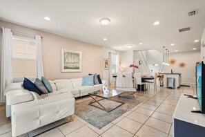 A modern living room with a white sofa, glass coffee table, and a dining area with white chairs. - EV_1800 - 6BR / 5BA Luxury Villa w/ Private Pool & Spa, Game Room, Resort Access (Kissimee)
