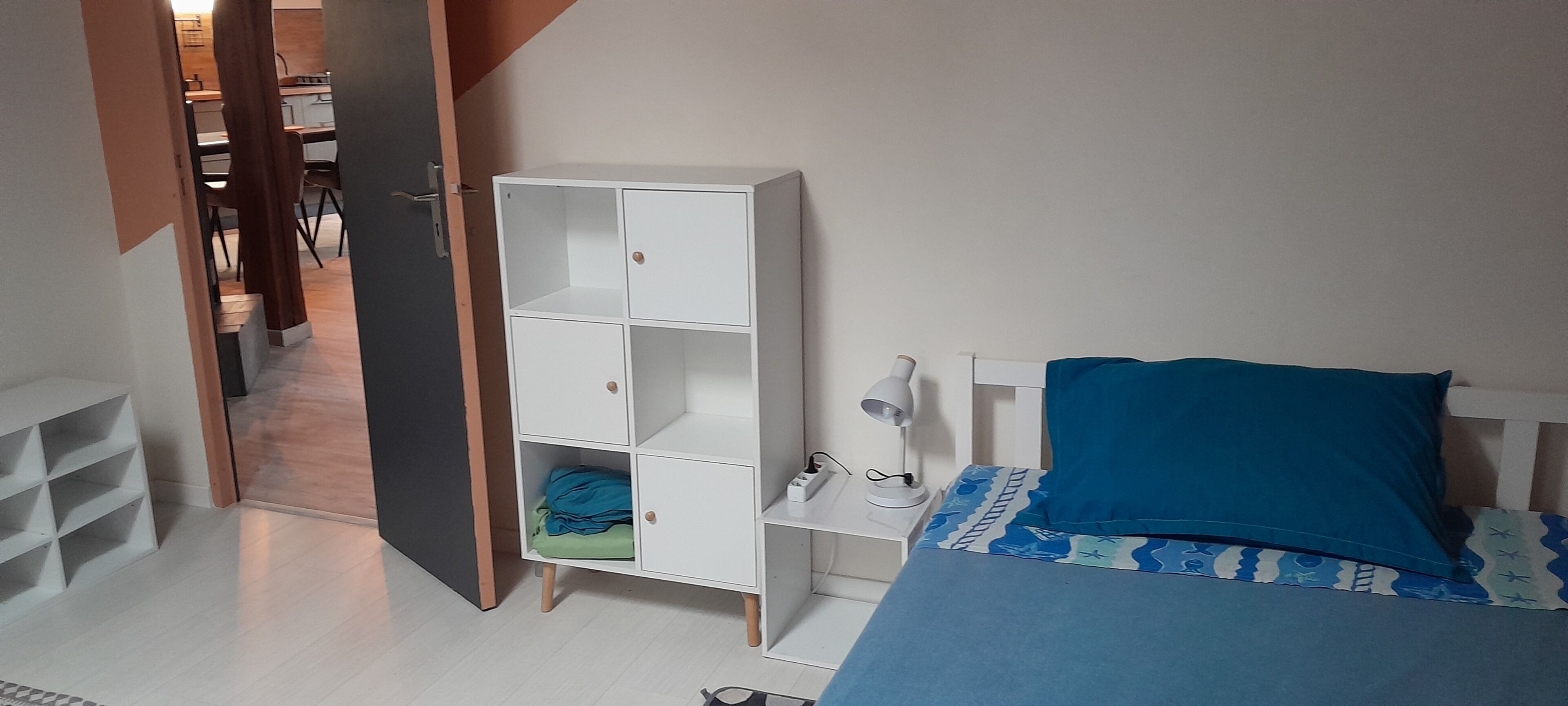 A bedroom with a bed, a white wardrobe, a desk with a lamp, and a door leading to a bathroom.