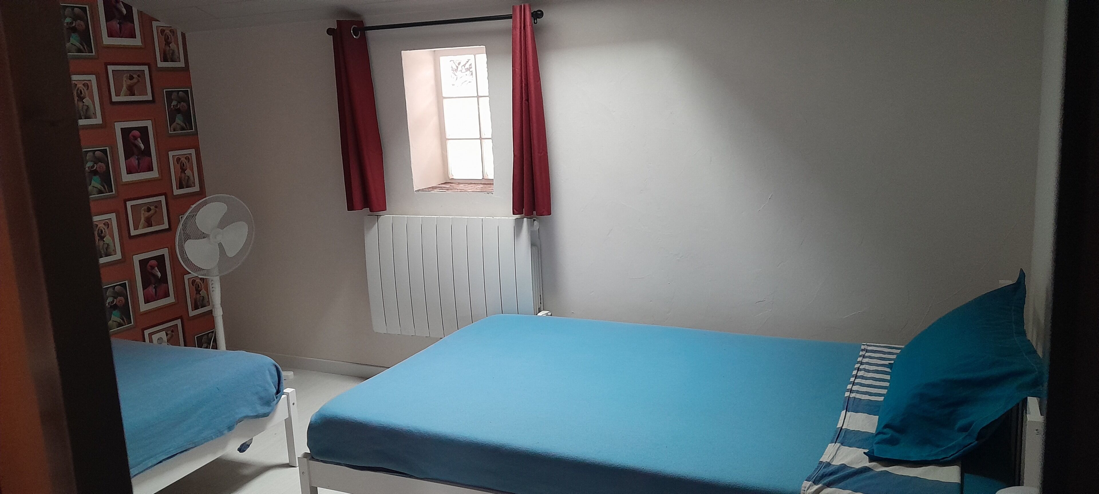 Una habitación con dos camas, una ventana con cortinas rojas, un radiador y un radiador y una pared con cuadros.