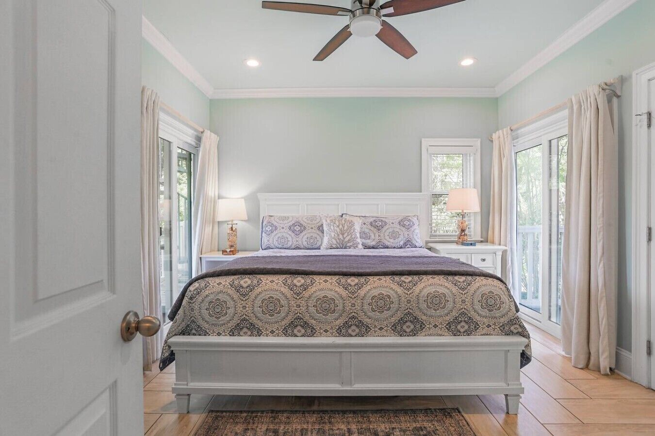 A bedroom with a bed, two bedside tables, a ceiling fan, and a view of the outdoors through windows.