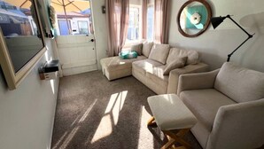 A cozy living room with a sectional sofa, a small ottoman, a TV, and a lamp.