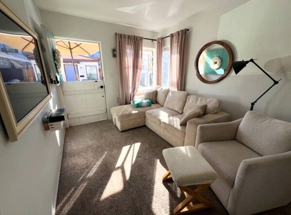 A cozy living room with a sectional sofa, a small ottoman, a TV, and a lamp.