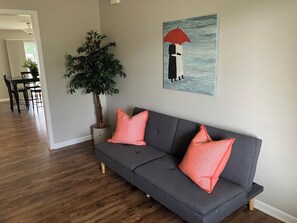 A gray sectional sofa with two pink pillows, a potted plant, and a painting of two figures under an umbrella. - Peaceful 3 bedroom handicapped accessible house with AC in charming Hamilton. (Hamilton)