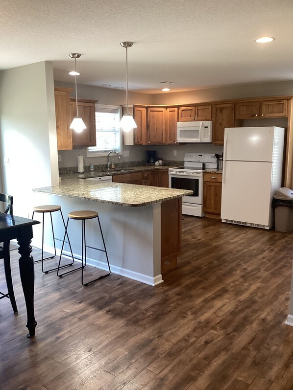 A kitchen with wooden cabinets, a white refrigerator, a microwave, a stove, and a granite countertop island with bar stools.
