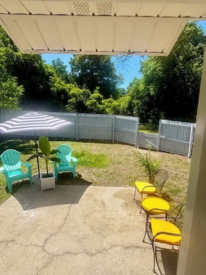 A patio area with a covered seating area, a white fence, and green foliage in the background. - Your Getaway Awaits (Pensacola)